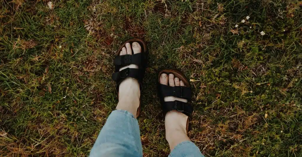 A person's feet wearing dark-colored sandals standing on grass.