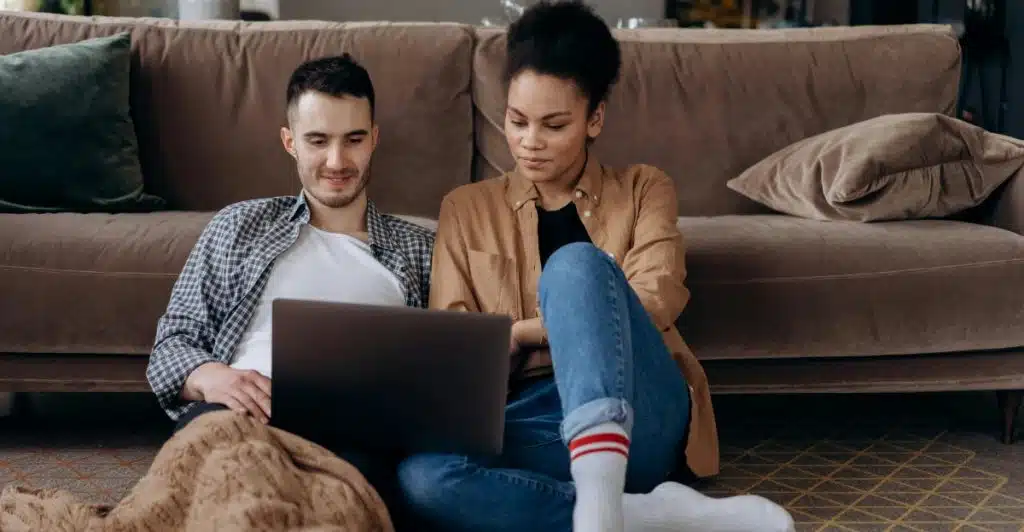 A couple sitting on the floor, looking at a laptop, with a blanket and pillows nearby.