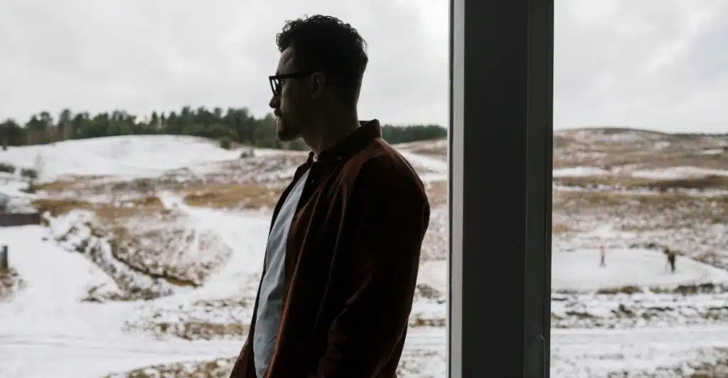 A man standing and looking out a window at a snowy landscape.