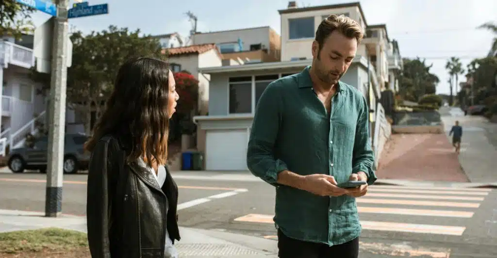  A man looking at his phone on a city street while a woman stands next to him, looking concerned.