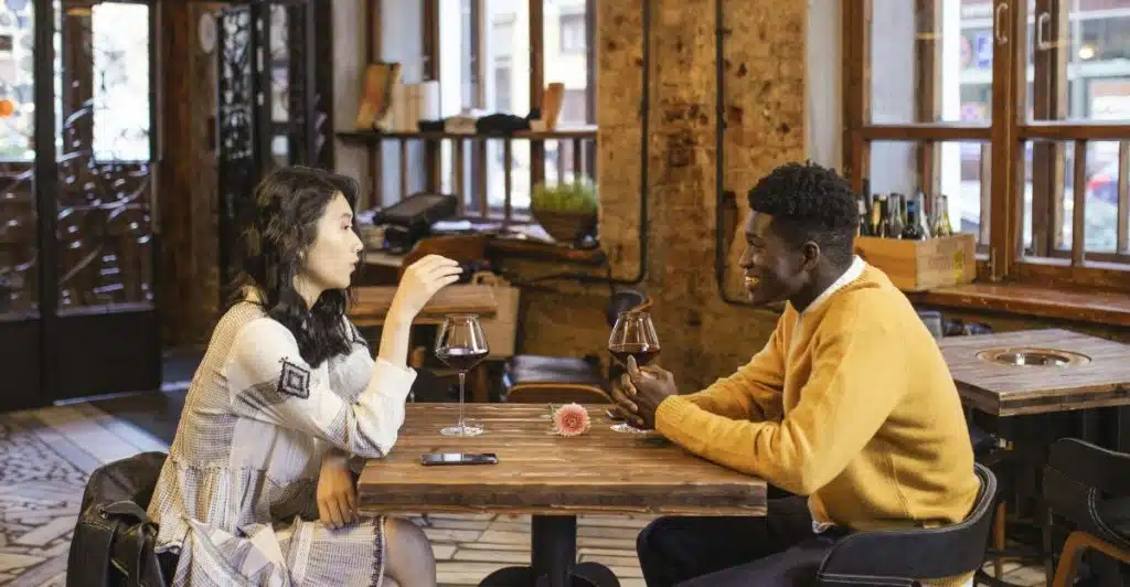 A man and a woman sitting at a table in a restaurant or cafe, talking and holding wine glasses.
