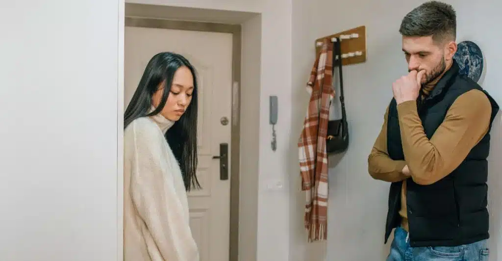 A man and a woman standing at a distance from each other in a hallway, both looking down.