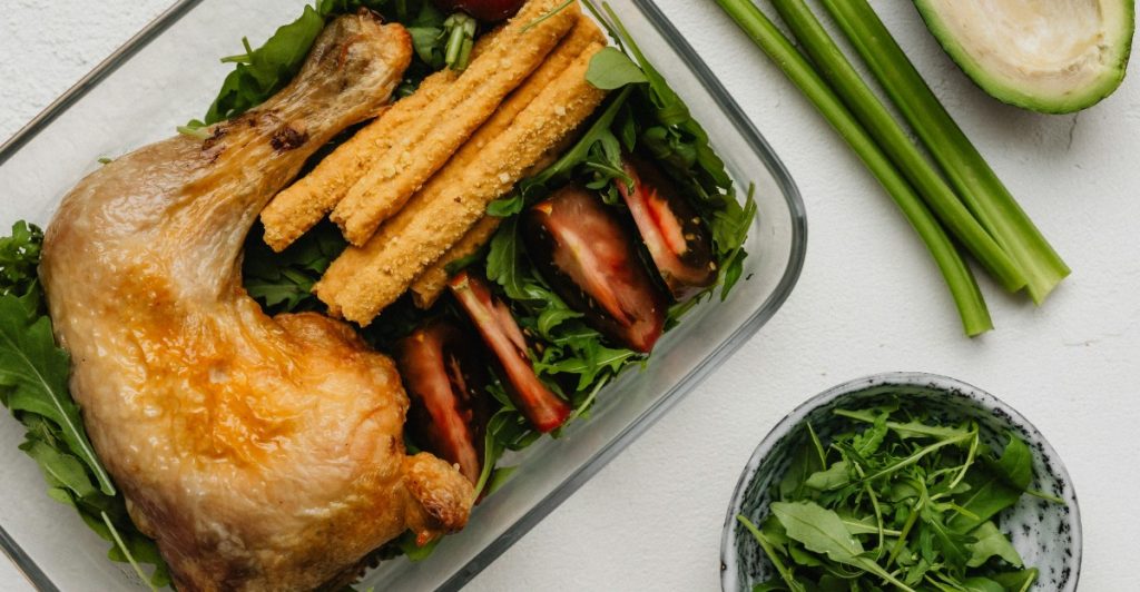 A meal prep container with a chicken leg, vegetables, and another dish with green leaves.