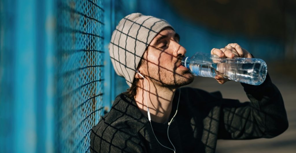 A person wearing a beanie and athletic wear drinking from a clear plastic water bottle while standing near a blue chain-link fence.