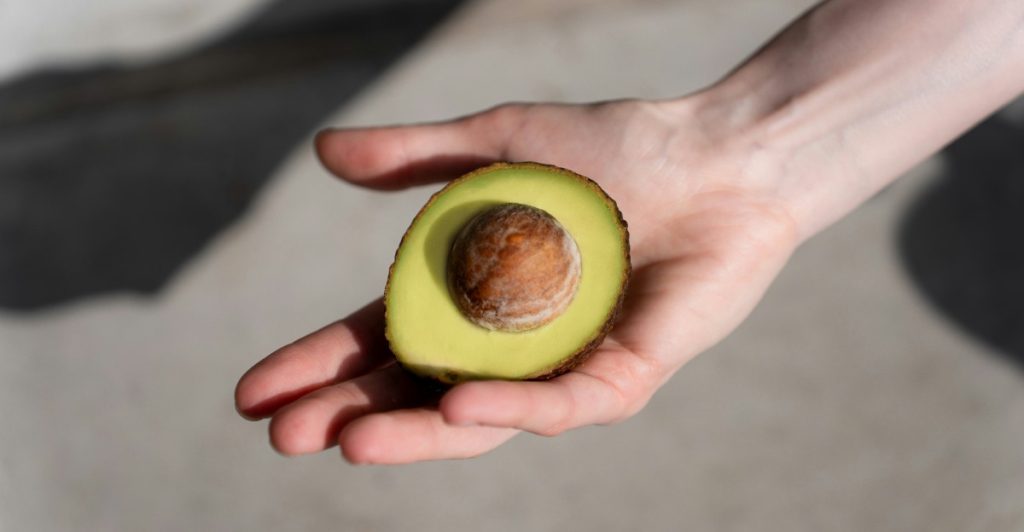 A hand holding half an avocado casting a shadow on the wall behind it.