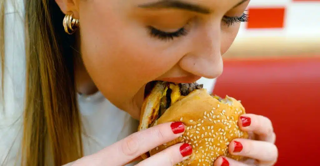 A person with red nail polish takes a large bite out of a sesame seed bun burger.