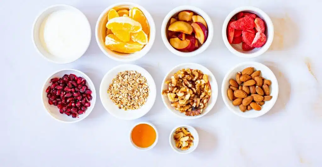 White bowls arranged on a white surface contain yogurt, sliced oranges, plums, freeze-dried strawberries, pomegranate seeds, granola, walnuts, almonds, and honey.