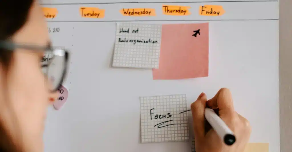 A person wearing glasses writing the word "Focus" on a sticky note attached to a white board covered in notes and days of the week.