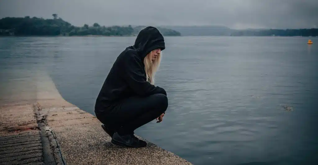 A woman with blonde hair wearing a black hoodie and dark clothes, crouching on a concrete edge next to a large body of water.