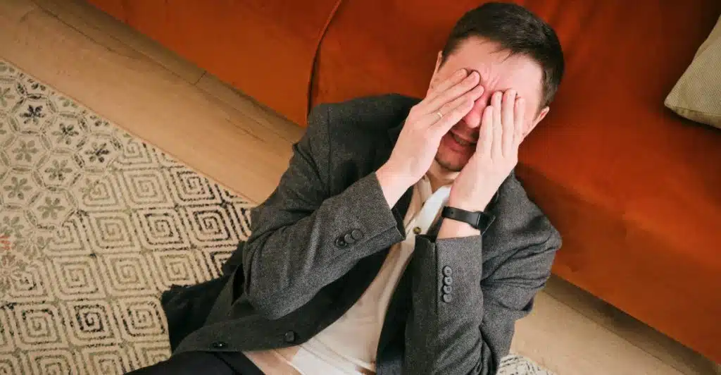 A man sitting on a rug with his hands covering his face, appearing distressed.
