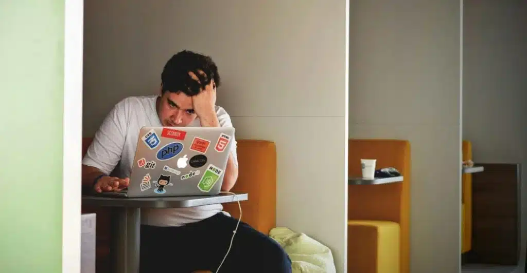 A person with dark hair wearing a white t-shirt with stickers is seated in a booth, looking stressed and holding their head while using a laptop.