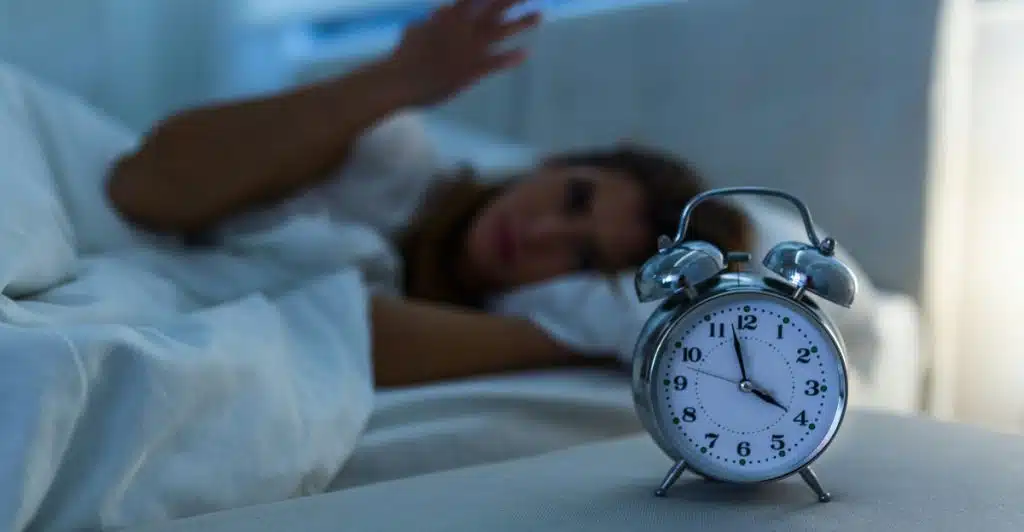 A silver alarm clock in the foreground on a bed, with a blurry image of a person struggling to sleep in the background.