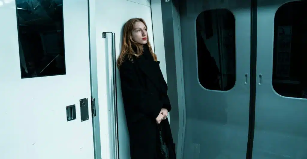 A woman in a black coat standing in a subway train, leaning against the wall and looking thoughtful.