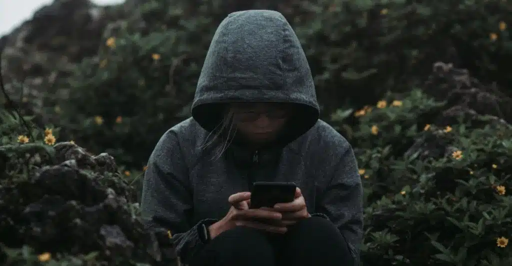A woman wearing a hooded jacket, looking down at her phone amidst green foliage and rocks.