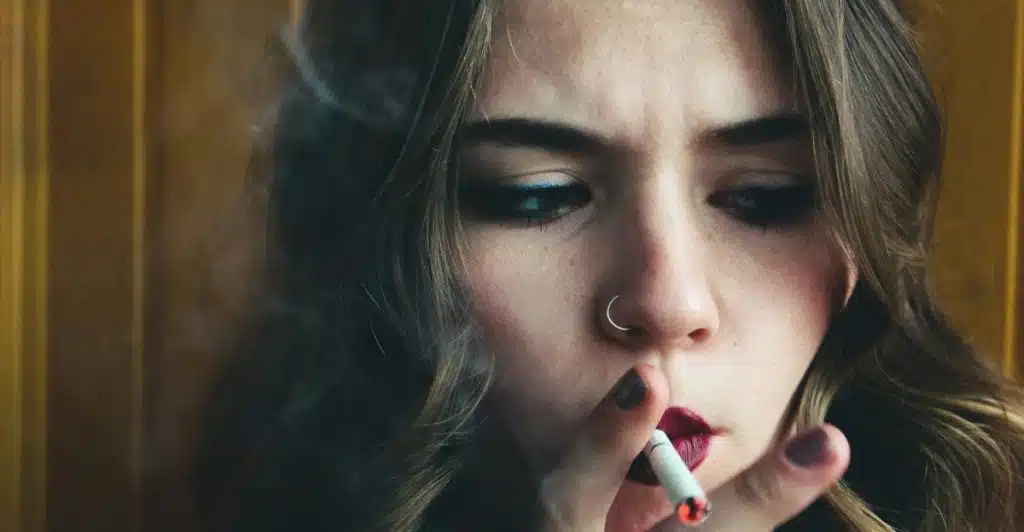 A woman with long hair and a nose ring, exhaling smoke from a cigarette she is holding.