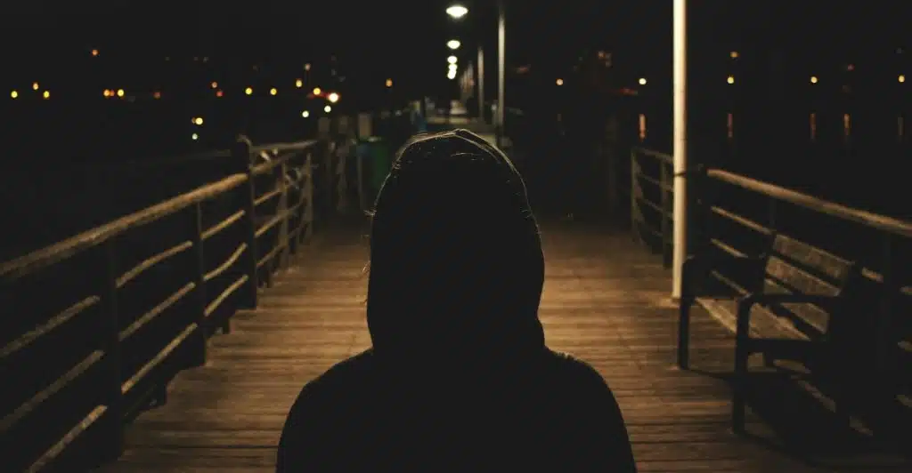 A silhouetted figure wearing a hooded top standing on a wooden pier at night, with distant lights.