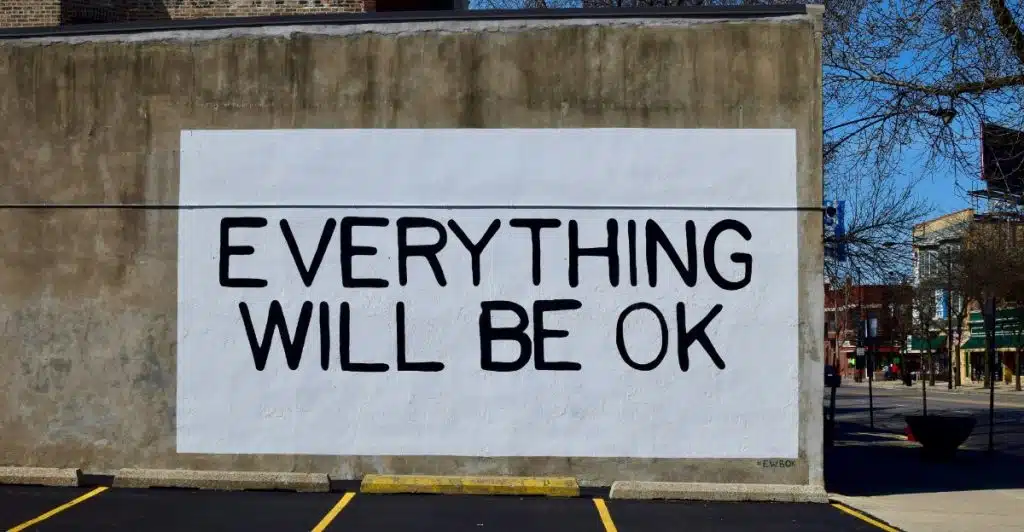 A mural on a concrete wall that reads "EVERYTHING WILL BE OK" in large black letters, with a parking lot in front.