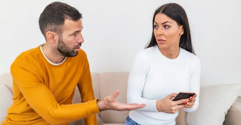 A man and a woman sitting on a couch, appearing to be in a disagreement, with the woman holding a phone.