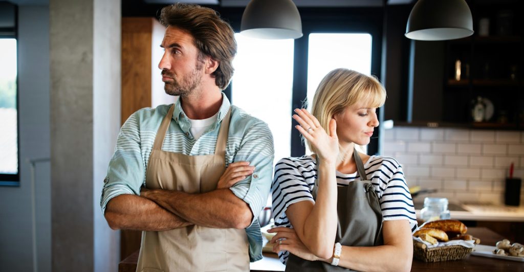 A man and a woman wearing aprons, standing with their arms crossed and backs to each other in a kitchen.