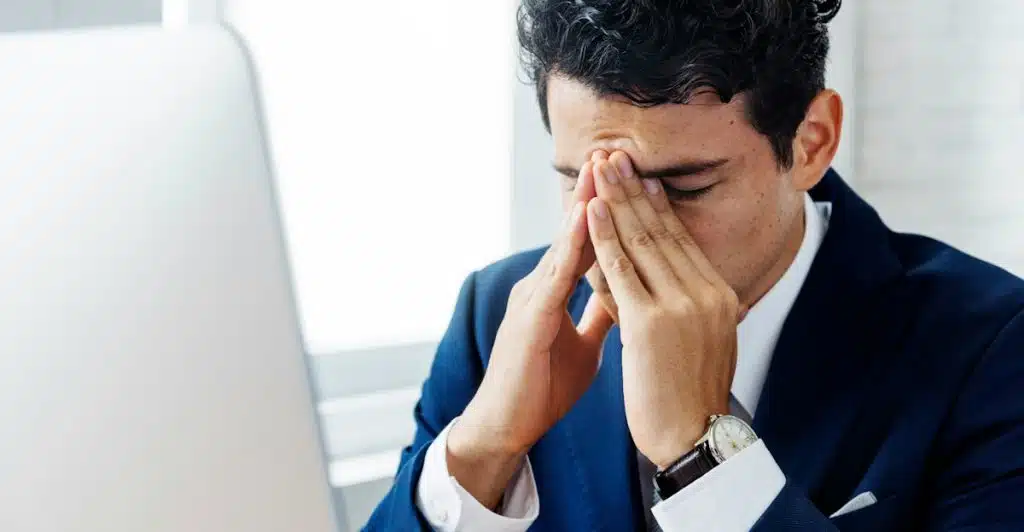 A man in a suit sitting at a desk with his head in his hands, appearing stressed.