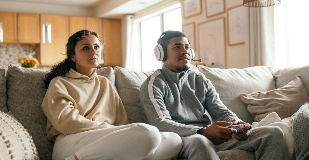A man and a woman sitting on a couch, with the man wearing headphones and playing a video game, and a small dog nearby.