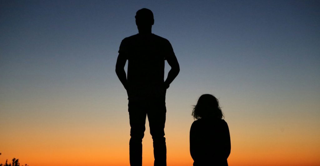 A silhouette of a tall man standing and a shorter woman sitting on a bench against a sunset.