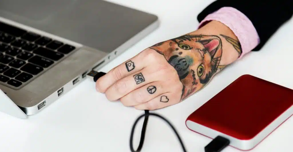 A person's tattooed hand plugging a USB cable from a red external hard drive into a silver laptop.