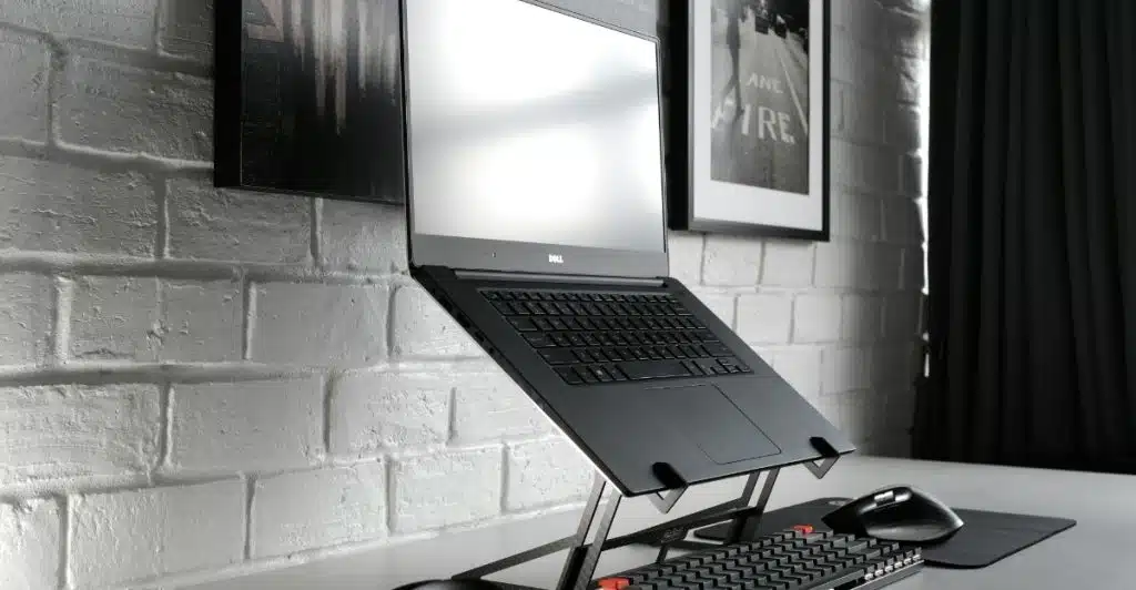 A black laptop elevated on a silver adjustable stand, with headphones and a keyboard on a white desk.