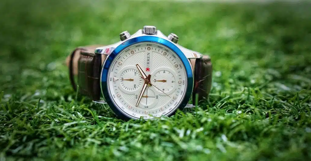 A close-up of a silver watch with a white face and brown leather strap, lying on vibrant green grass.
