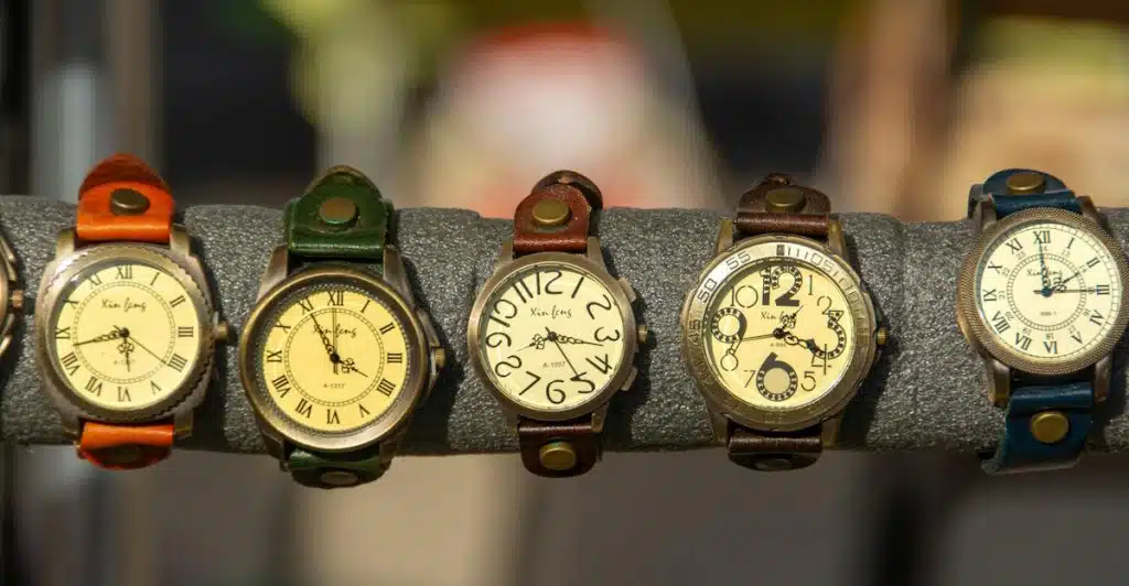 A row of five vintage-style watches with various colored leather straps and light-colored dials, displayed on a grey stand.