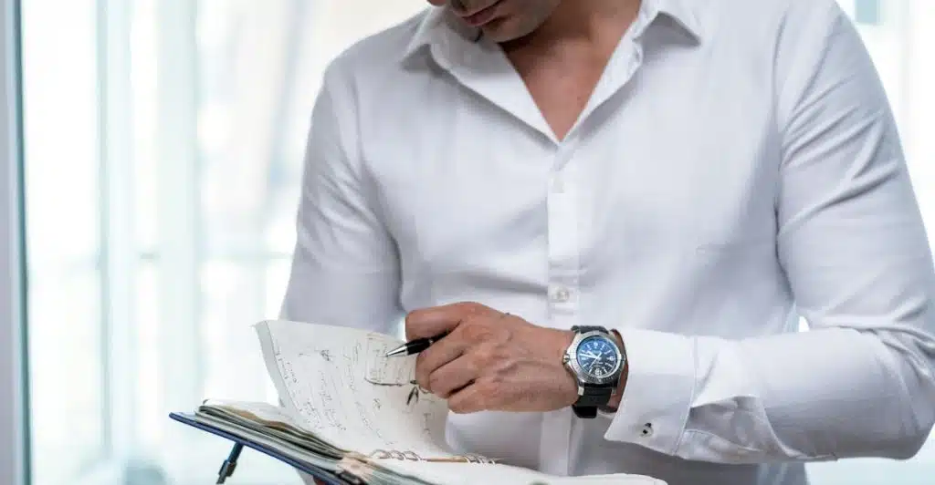 A man in a white long-sleeve shirt and khaki pants, wearing a watch, holding a notebook and writing with a pen.