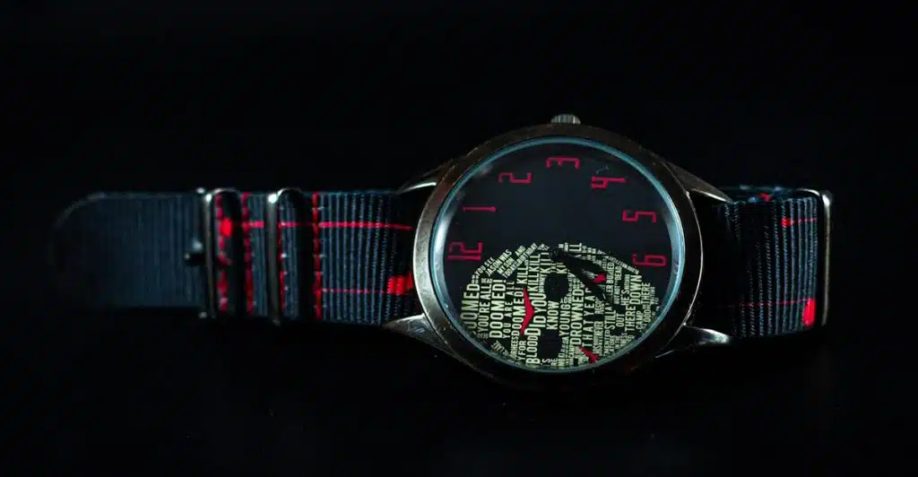 A close-up of a dark-faced watch with a red and black striped NATO strap, featuring a skull design made of text on the dial.
