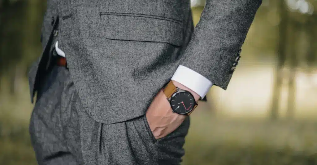 A man wearing a grey suit jacket and a brown leather watch, with his hand in his pocket.