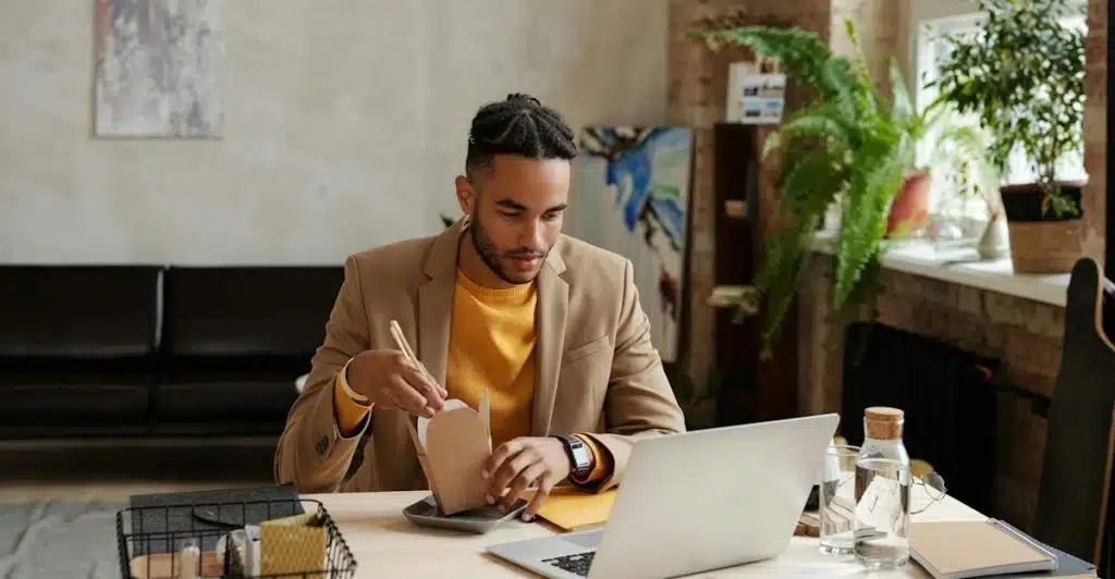 A man with dreadlocks and a light brown suit jacket over a yellow shirt, sitting at a desk with a laptop, eating noodles from a takeout box.