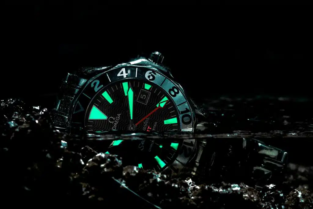 A close-up of a silver watch submerged partially in dark water, with its luminous hands and markers glowing green.