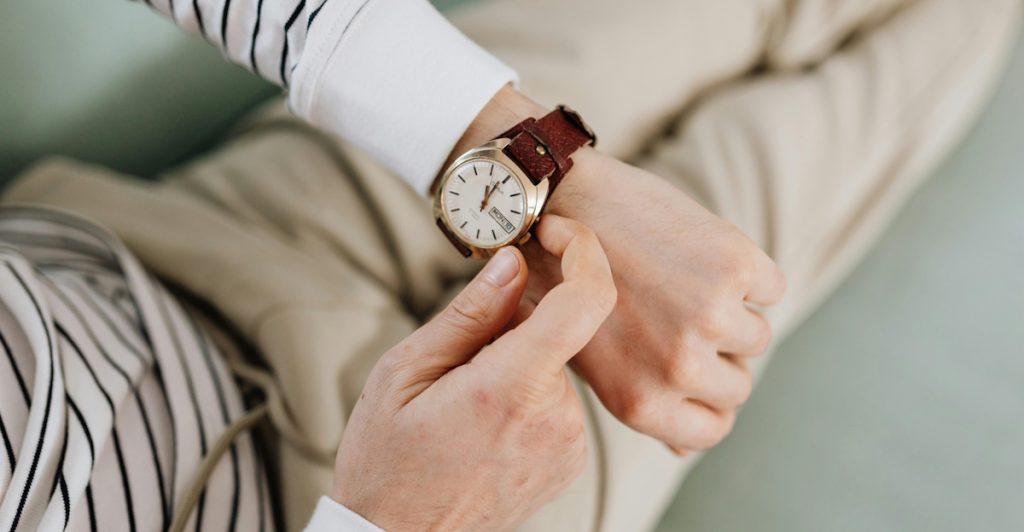 A man wearing a striped shirt and khaki pants, looking down at a vintage-style watch with a brown strap on his wrist.
