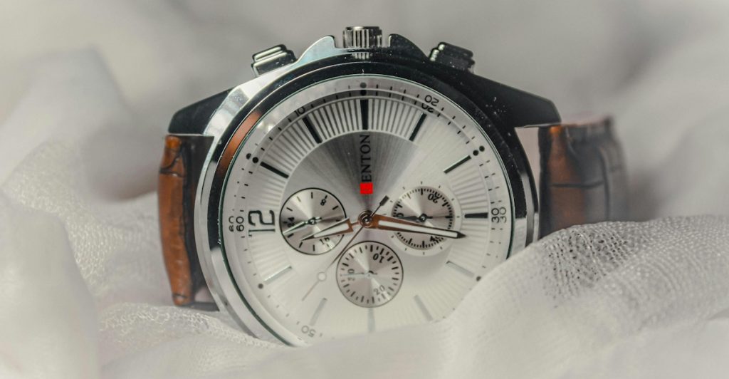 A close-up of a polished wristwatch with a white dial and brown leather strap, resting on white fabric.