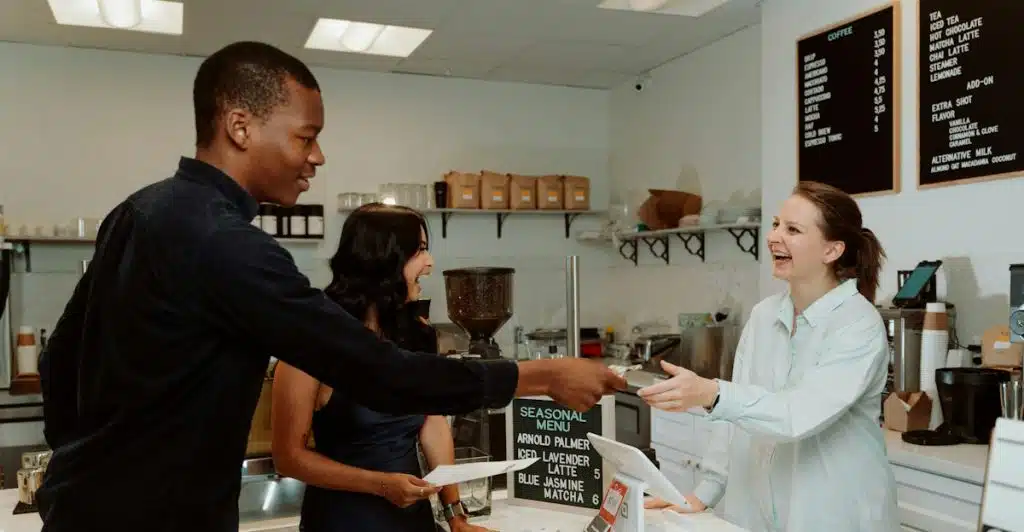 A man handing a card to a barista at a coffee shop counter, with a woman standing beside him looking towards the barista.