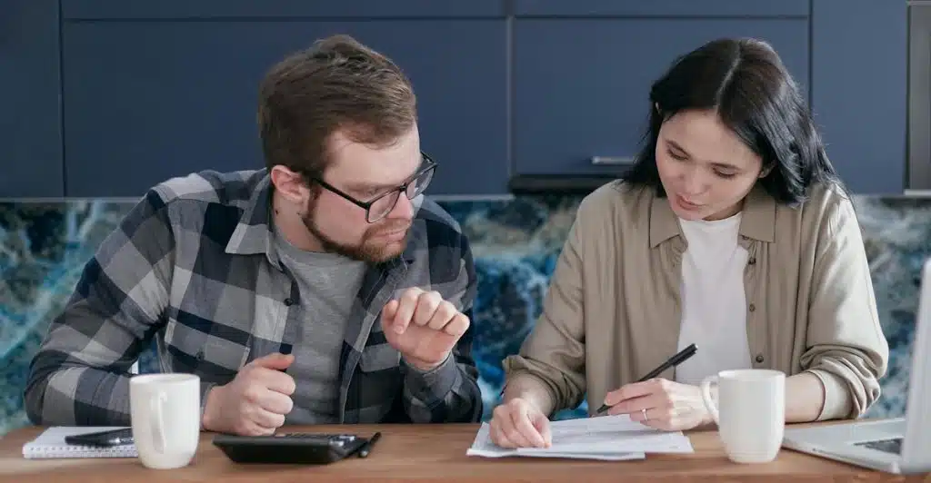 A man and a woman sitting at a wooden table in a kitchen looking at papers and using a calculator, with mugs and a laptop also on the table.