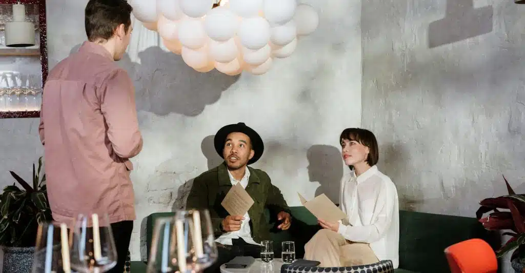 Two people seated at a table in a restaurant looking at menus, while a third person stands beside their table talking to them.