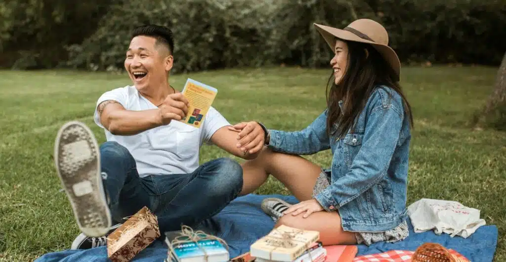 A man and a woman sit together on a blue blanket on the grass outdoors, laughing while holding a book or card, with books and fruit around them.