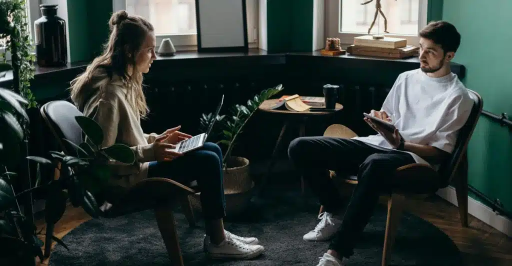 A woman sitting in a chair with a laptop looking at a man who is sitting opposite her, holding a tablet, with plants and a lamp in the background.