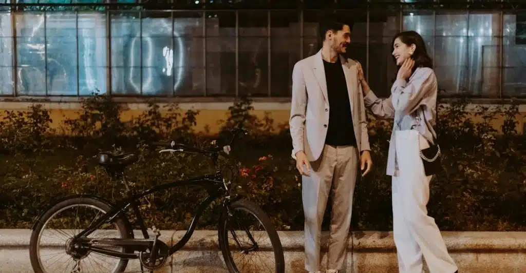 A man and a woman standing on a paved area at night next to a bicycle, with the man wearing a light-colored suit and the woman touching his arm and smiling at him.