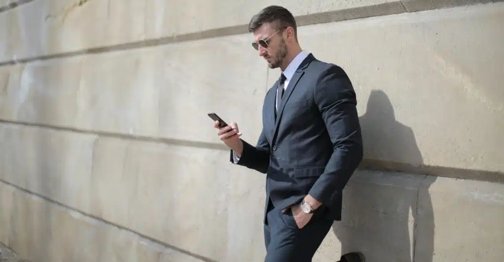 A man in a suit and sunglasses standing against a concrete wall, looking at his phone.