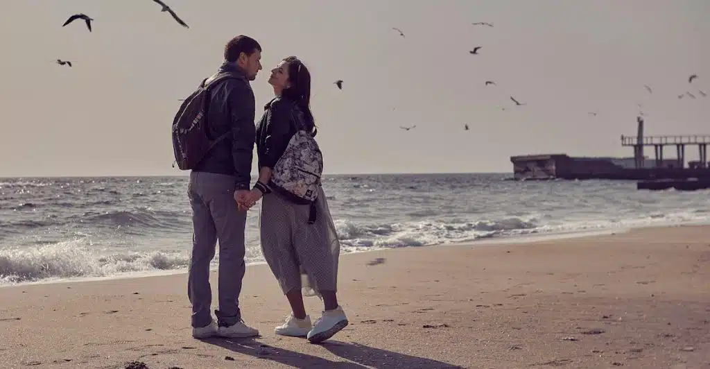 A couple standing on a sandy beach, holding hands and looking at each other with birds flying in the sky.