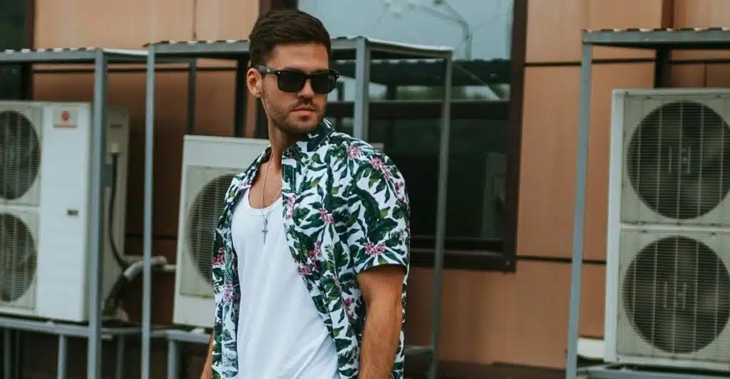 A man in a white floral short-sleeve shirt, white t-shirt, and jean shorts with sunglasses looking to the side.