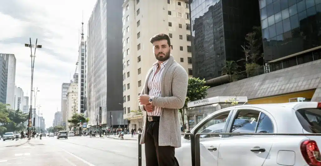 A man in a striped shirt and long grey cardigan standing on a city street.