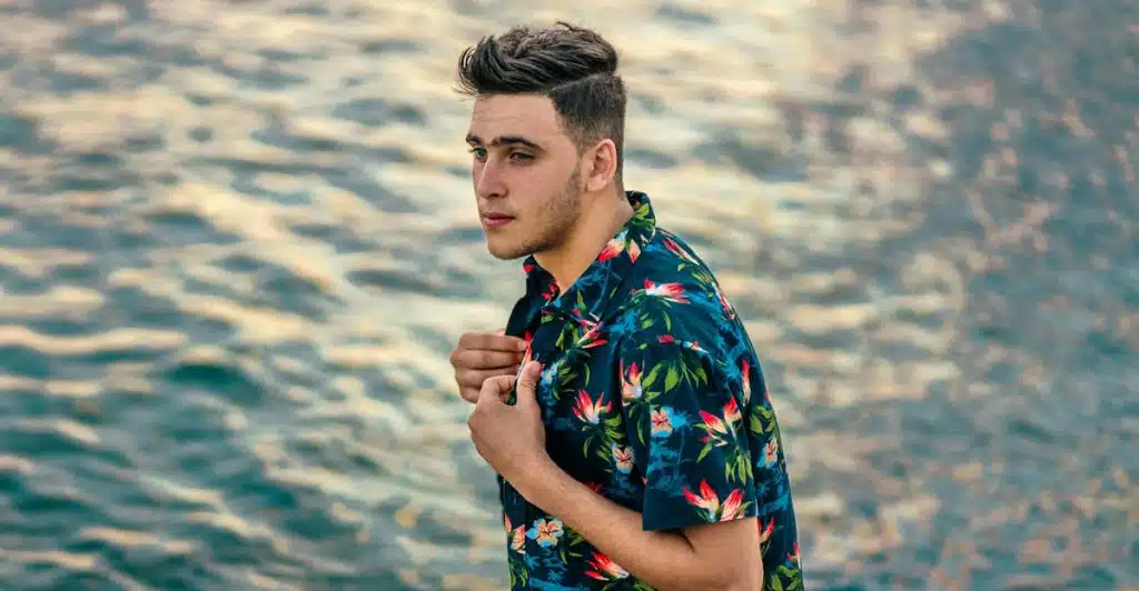 A man in a dark blue floral printed short-sleeve shirt and leopard print shorts by the water.
