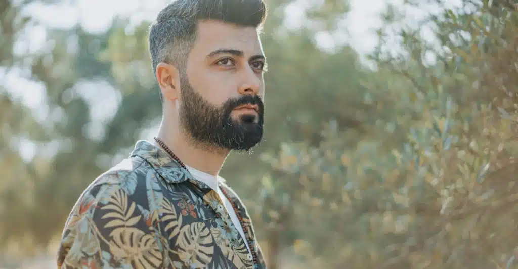 A man with a beard and a tropical printed shirt standing outdoors among trees.