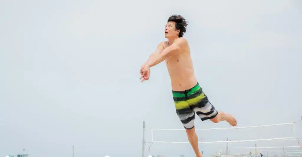 A man in mid-air wearing striped swim trunks on a sandy beach with a volleyball net in the background.